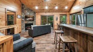 A cabin interior with a stone fireplace, wooden walls, a ceiling fan, and a kitchen area.