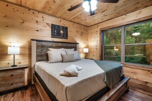 Een slaapkamer in een houten blokhut met een bed, nachtkastjes en een raam met uitzicht op bomen.