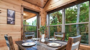 A wooden dining table set with plates, glasses, and a vase, surrounded by chairs, with large windows offering a view of trees and a mountain.
