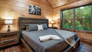 A wooden cabin bedroom with a large bed, two bedside tables with lamps, a view of the forest through a window, and a painting on the wall.