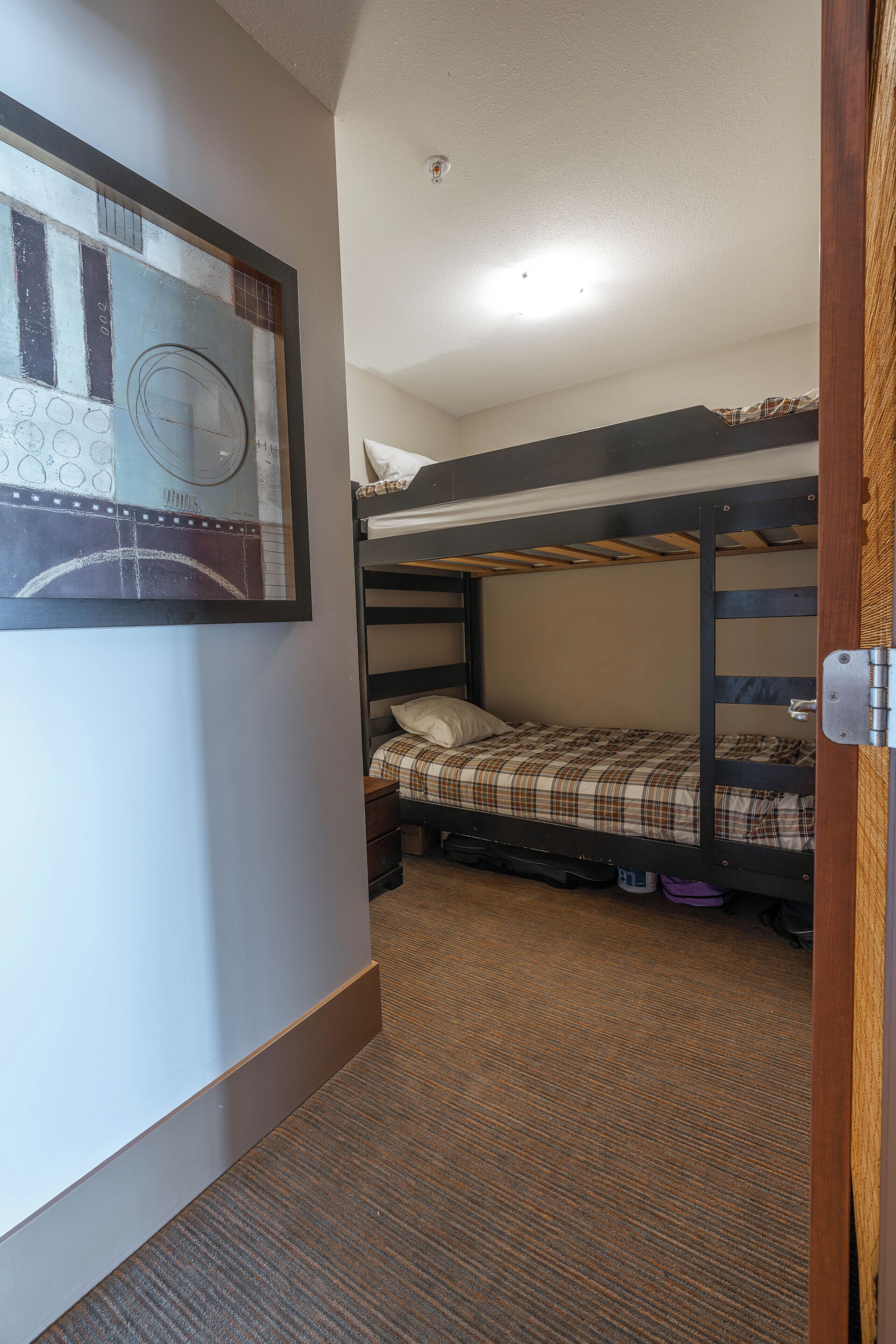 Une chambre avec des lits superposés, un tableau abstrait encadré accroché au mur, un sol recouvert de moquette et un chambranle de porte visible.