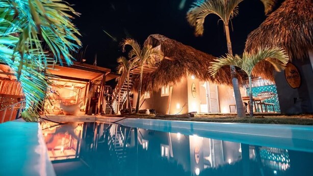 Zona junto a la piscina con un edificio de techo de paja, palmeras y una piscina cristalina que refleja el entorno.