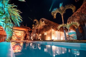 A poolside area with a thatched-roof building, palm trees, and a clear pool reflecting the surroundings. - KOKOHAU BUNGALOWS (Santa Verónica)