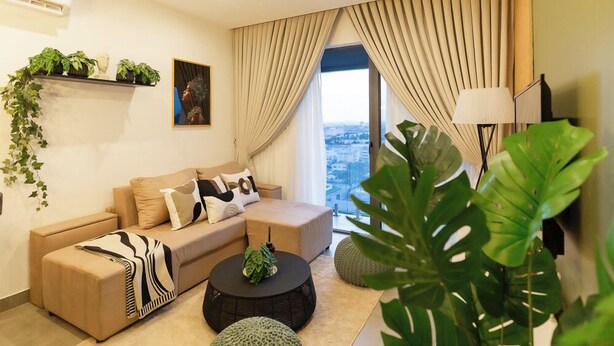 A modern living room with a sofa, ottoman, and coffee table, a large window with curtains, and a view of a cityscape. - NOVA-BlueWaters Property Management (Accra)
