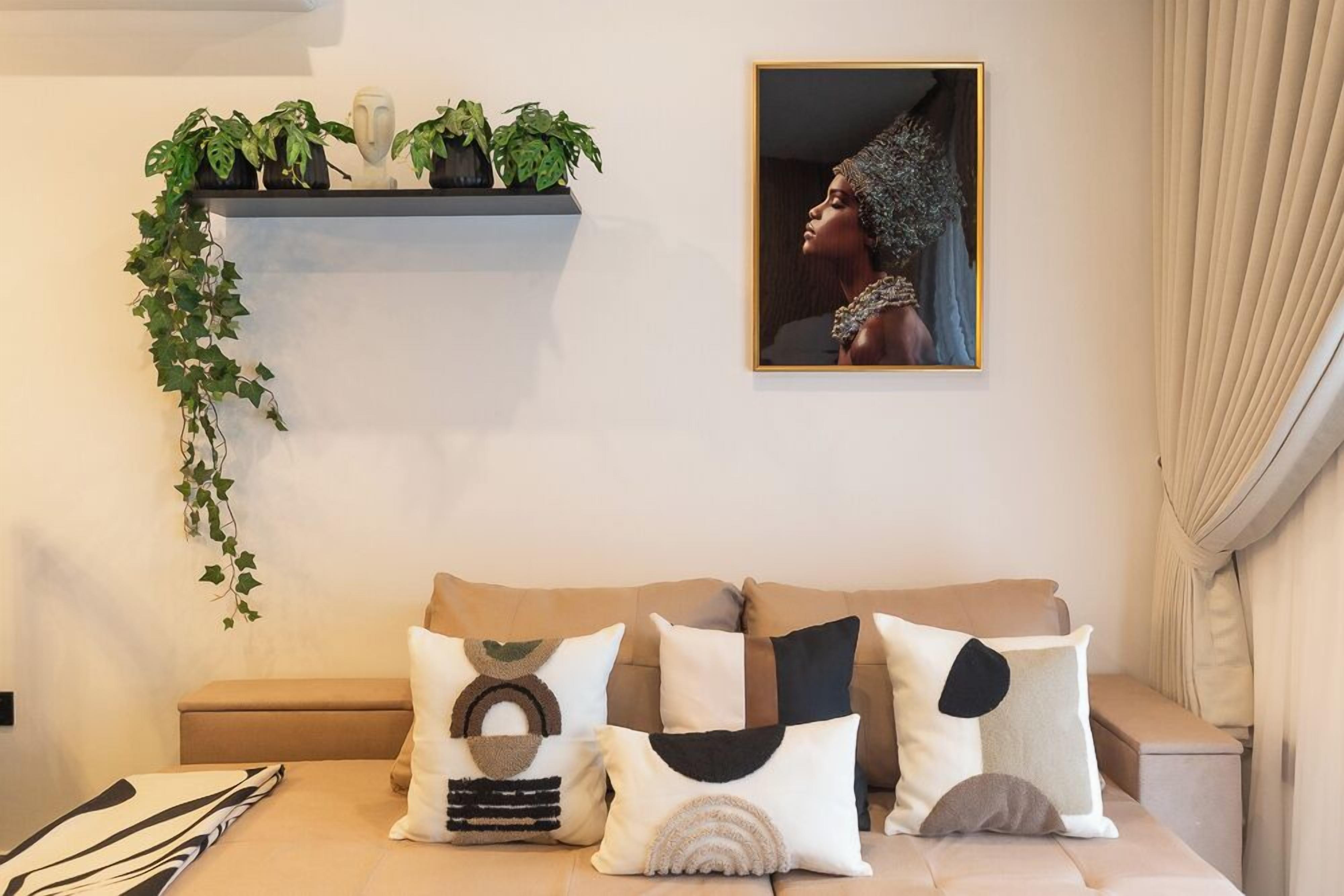 Un salón con un sofá decorado con cojines estampados, una estantería en la pared con plantas y una obra de arte enmarcada.