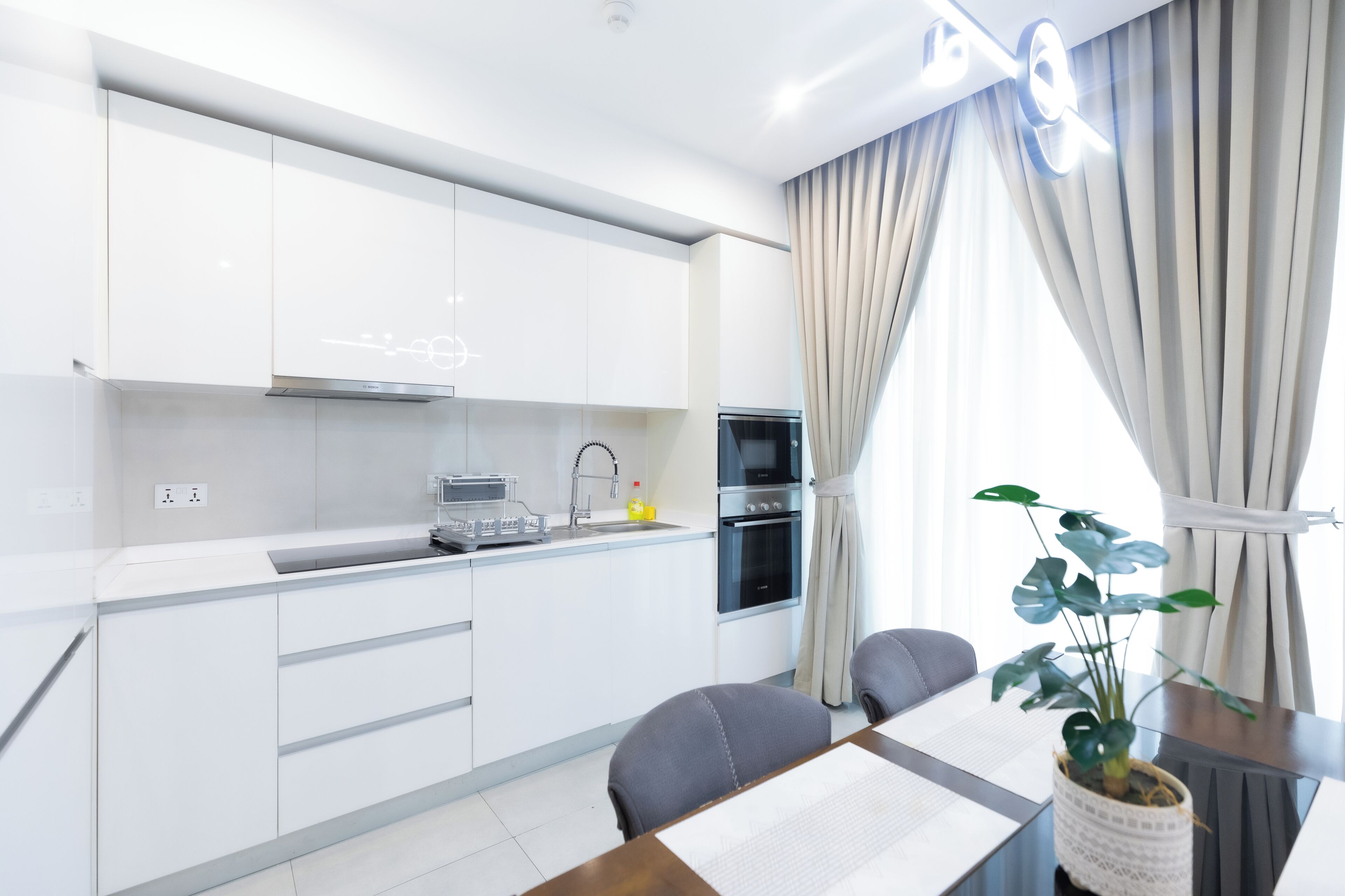 Een moderne keuken met witte kasten, een Bosch vaatwasser en een oven. Er is een eethoek met een tafel en stoelen, en een plant op de tafel.