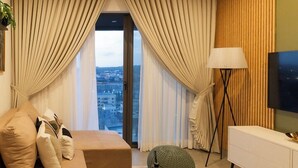 A modern living room with a sofa, a floor lamp, a TV, and a view of the city through large windows. - NOVA-BlueWaters Property Management (Accra)
