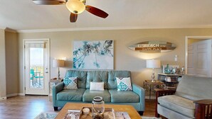 A living room with a teal sofa, a wooden coffee table with decorative items, a ceiling fan, and a wall-mounted sign. - Shared Pool, Hot Tub, Pet Friendly (Port Aransas)