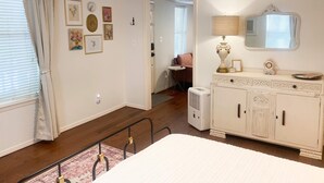 Un dormitorio con cama, mesita de noche, lámpara, cómoda y espejo. Hay una puerta que da a otra habitación.