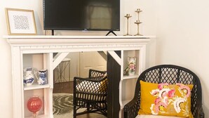 Una sala de estar con chimenea blanca, un televisor negro encima, un sillón de mimbre negro con un cojín amarillo y varios objetos decorativos en estanterías.