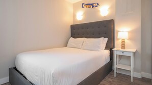 Ein ordentlich bezogenes Bett mit grauem Kopfteil, ein Nachttisch mit Lampe und eine Wandleuchte.