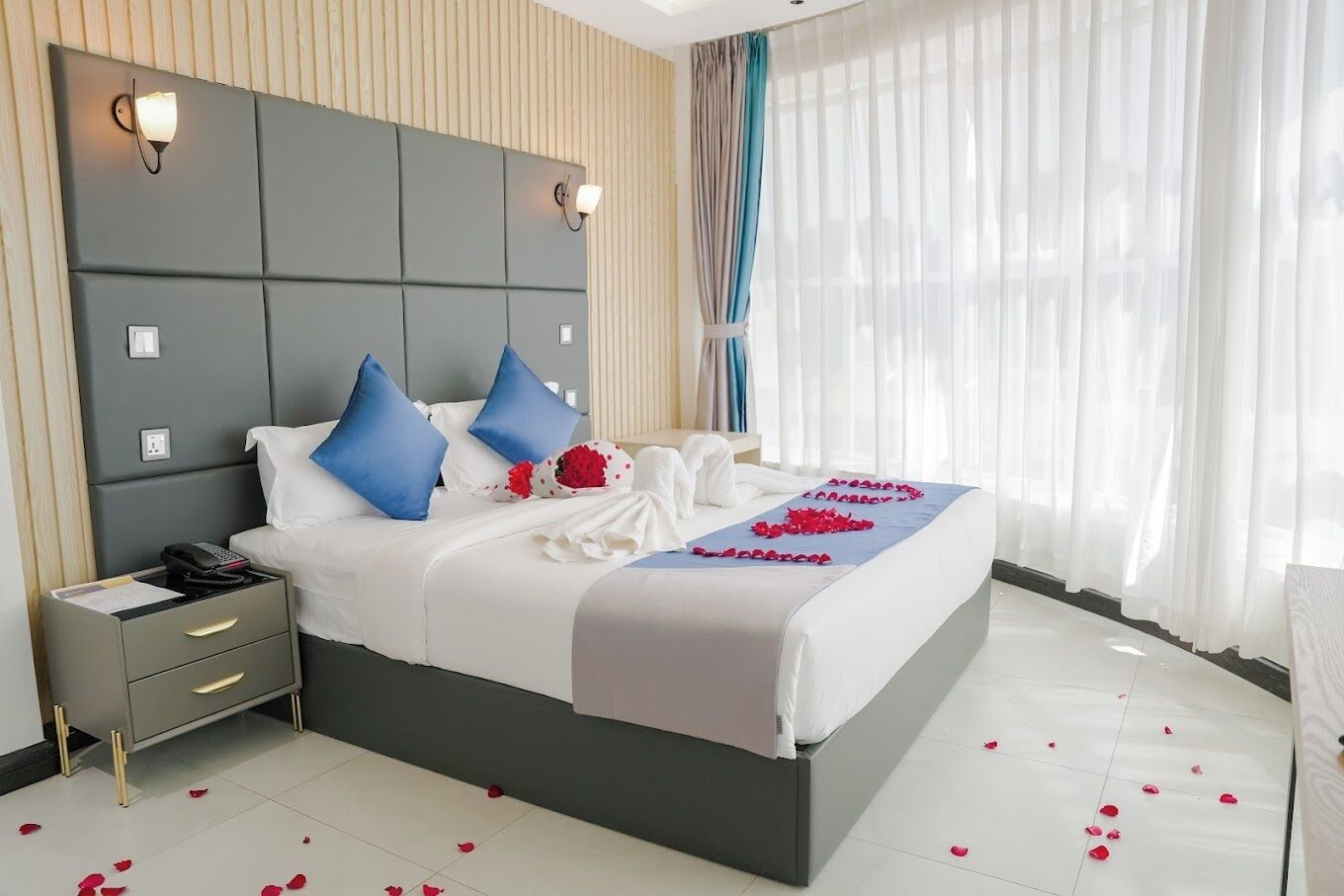 Quarto de hotel moderno com uma cama grande, mesas de cabeceira e vista para a cidade através de cortinas leves.