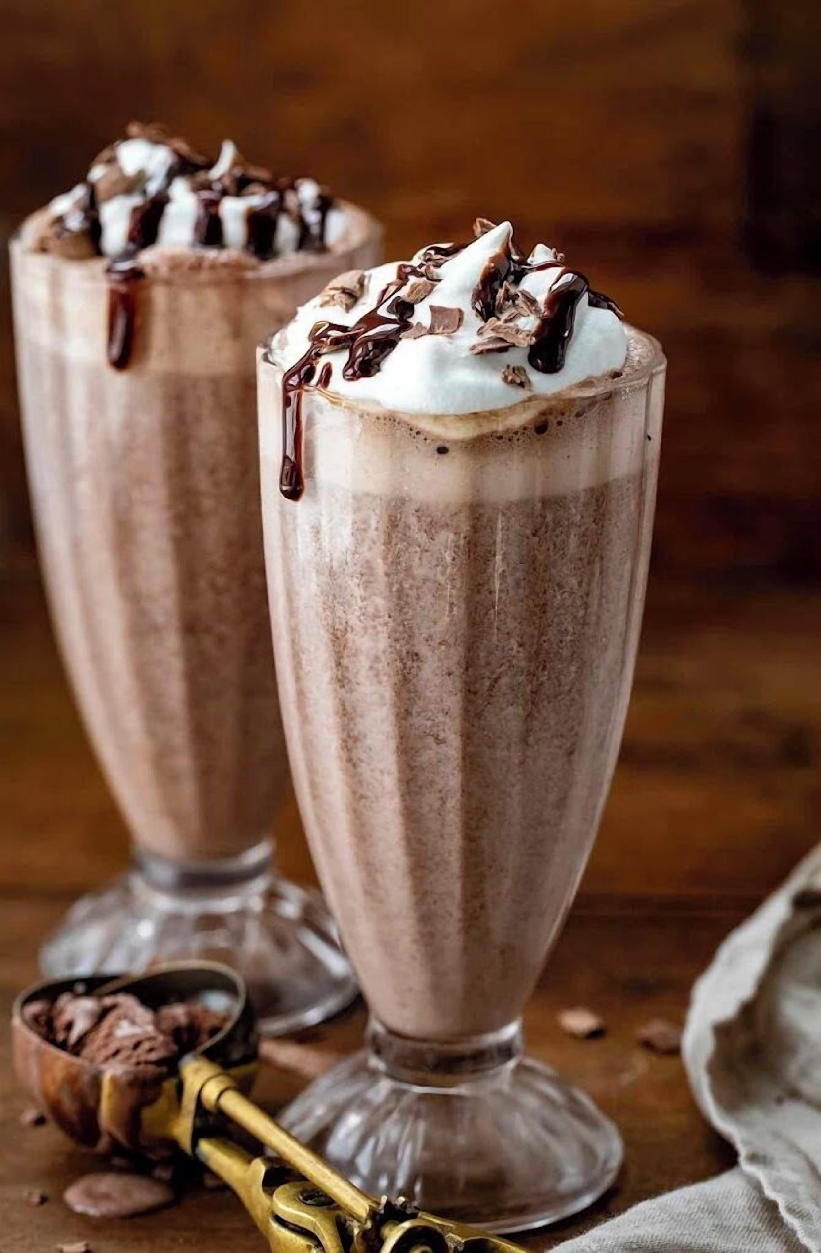 Two tall glasses of chocolate milkshakes with whipped cream and chocolate drizzle.