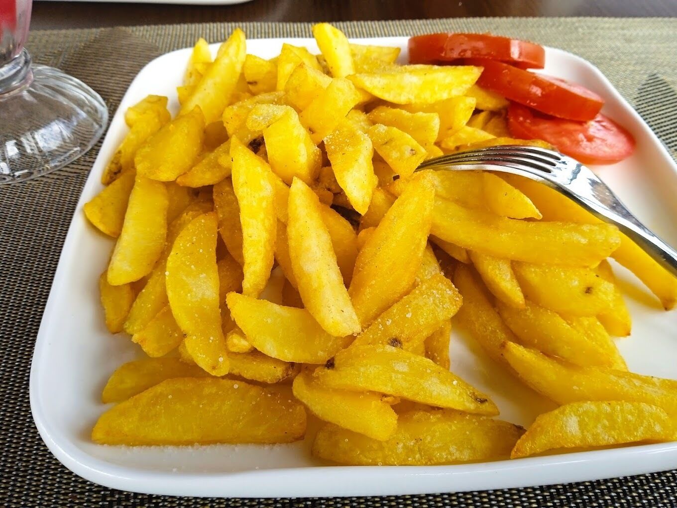 Un plato de papas fritas doradas, un tenedor y tomates en rodajas sobre una mesa.