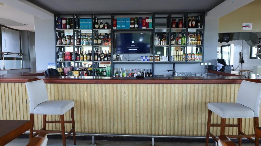 Un espace bar avec un comptoir en bois, deux tabourets de bar blancs rembourrés et des étagères remplies de différentes bouteilles et de verres.