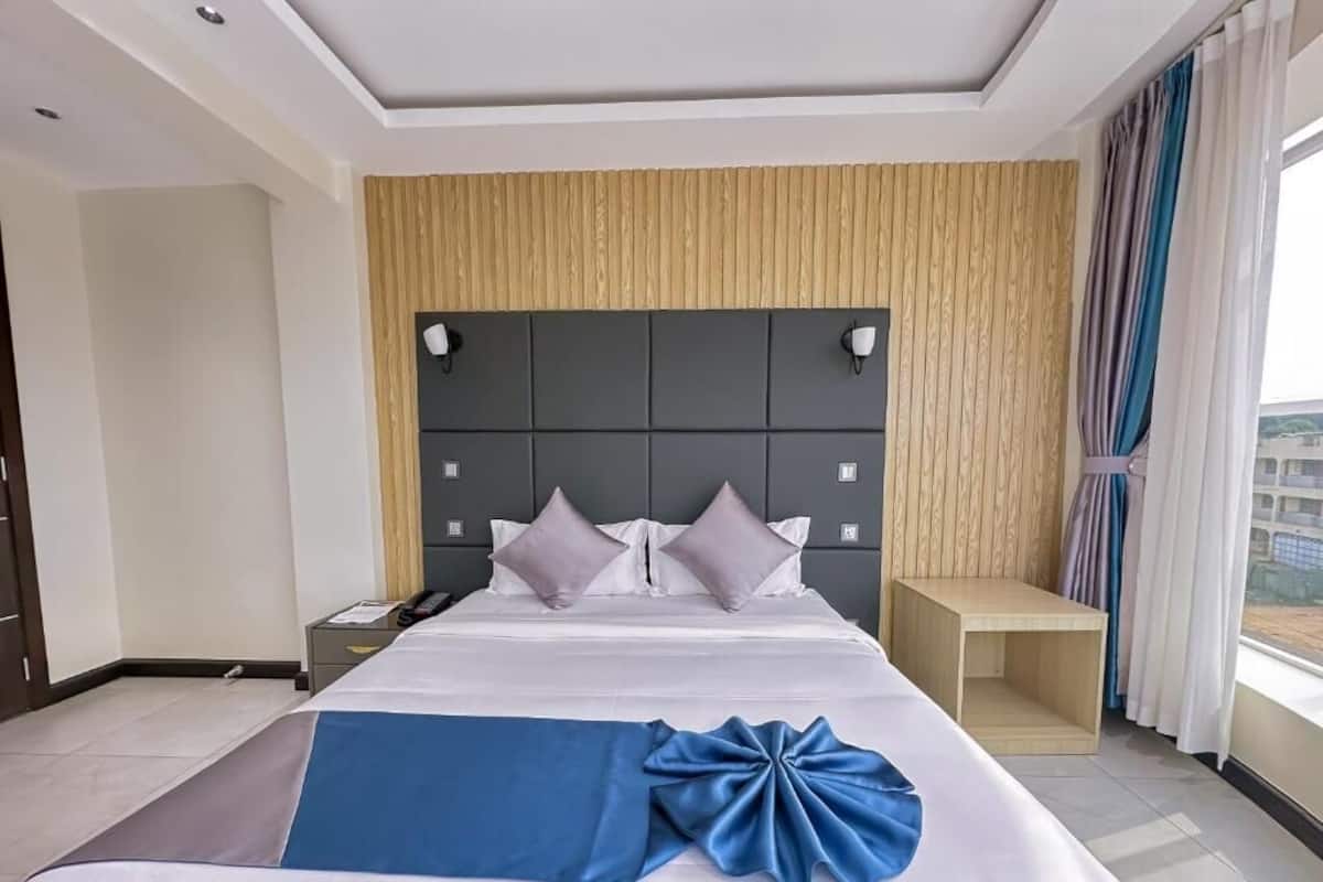 A hotel room with a large bed, a wooden headboard, a bedside table, and a view of the outside through a window.
