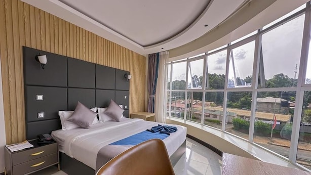 A modern hotel room with a large bed, a wooden headboard, a nightstand, a desk, and a large window offering a city view. - Zuri Place Hotel (Oyugis)