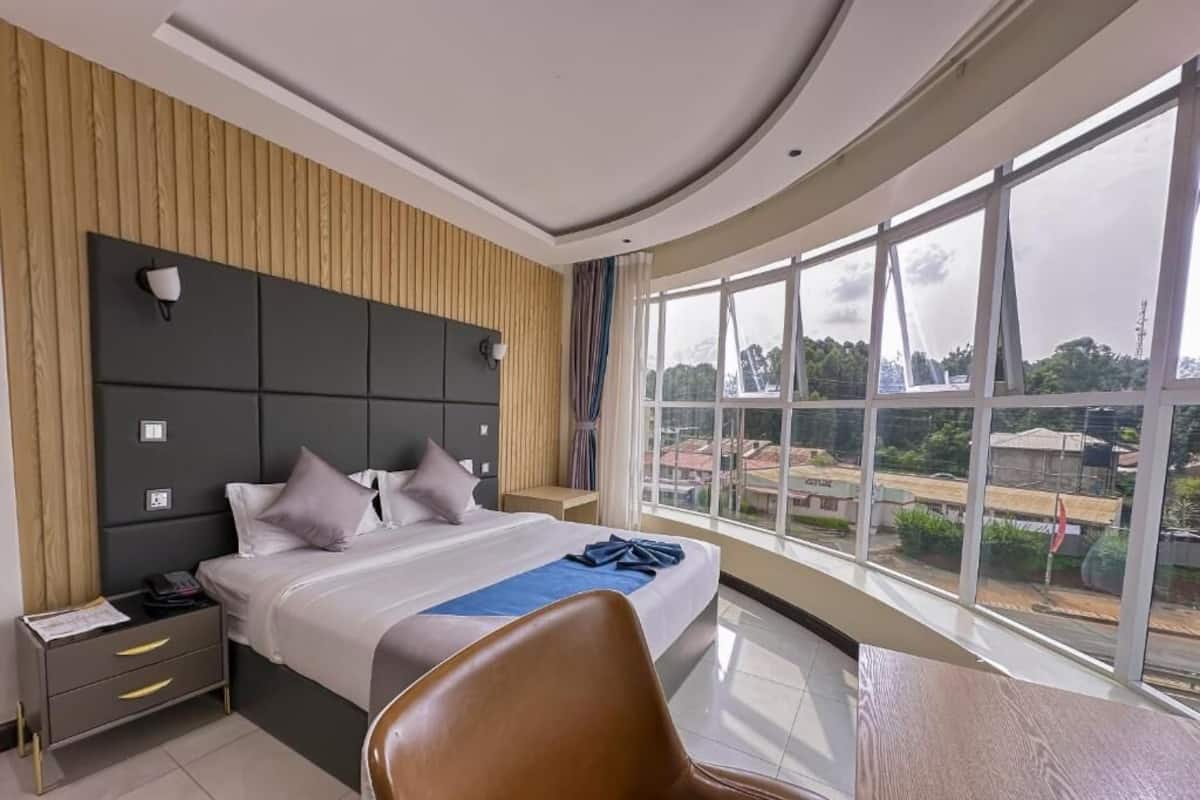 A modern hotel room with a large bed, a wooden headboard, a nightstand, a desk, and a large window offering a city view.