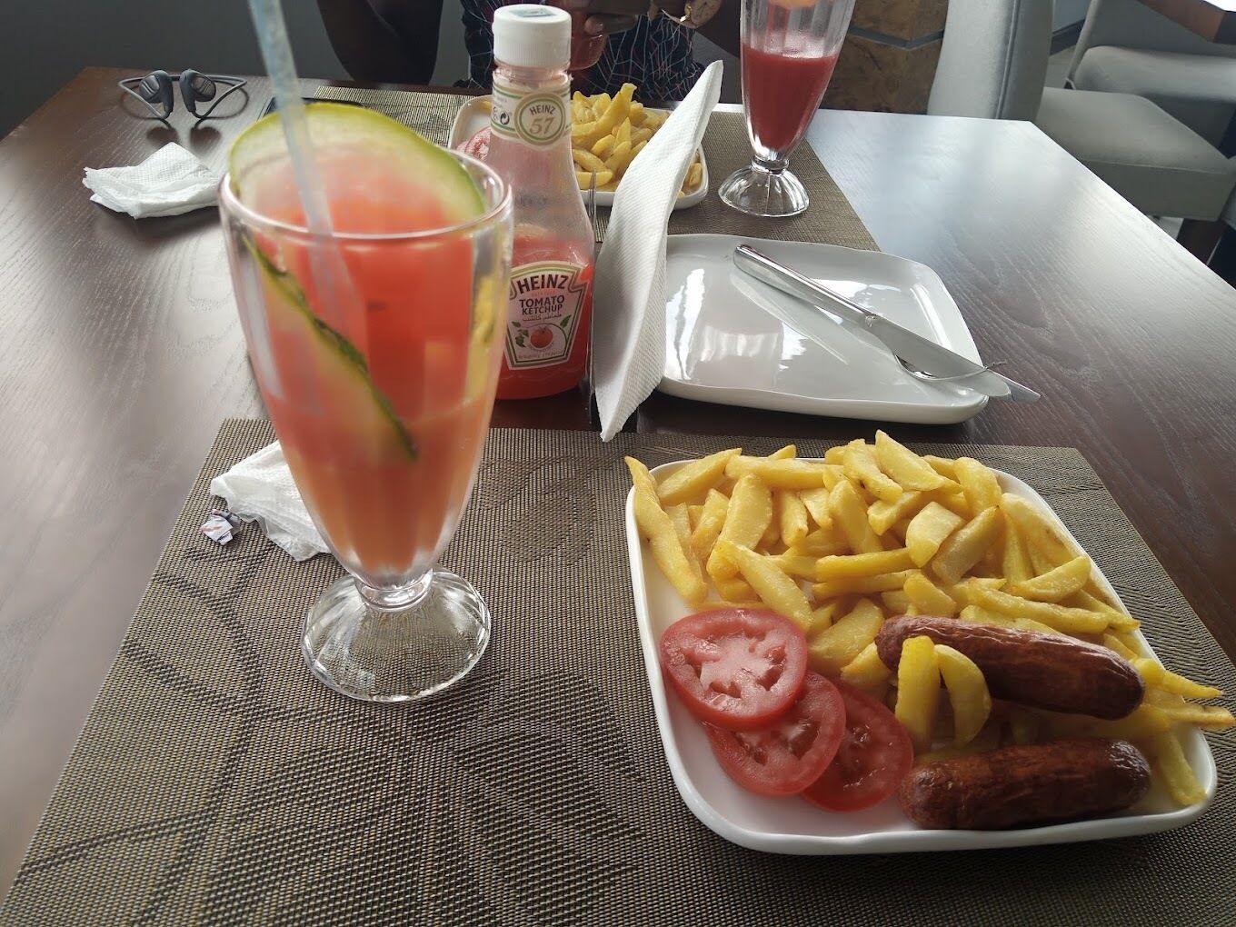 Un tavolo con un piatto di patatine fritte, salsicce e pomodori a fette, un bicchiere di succo di pomodoro e una bottiglia di Heinz ketchup.