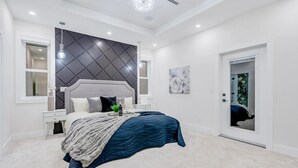 Une chambre à coucher avec un grand lit, une tête de lit moderne, une table de chevet, une commode, un luminaire fixé au mur et une porte dotée d’un miroir.