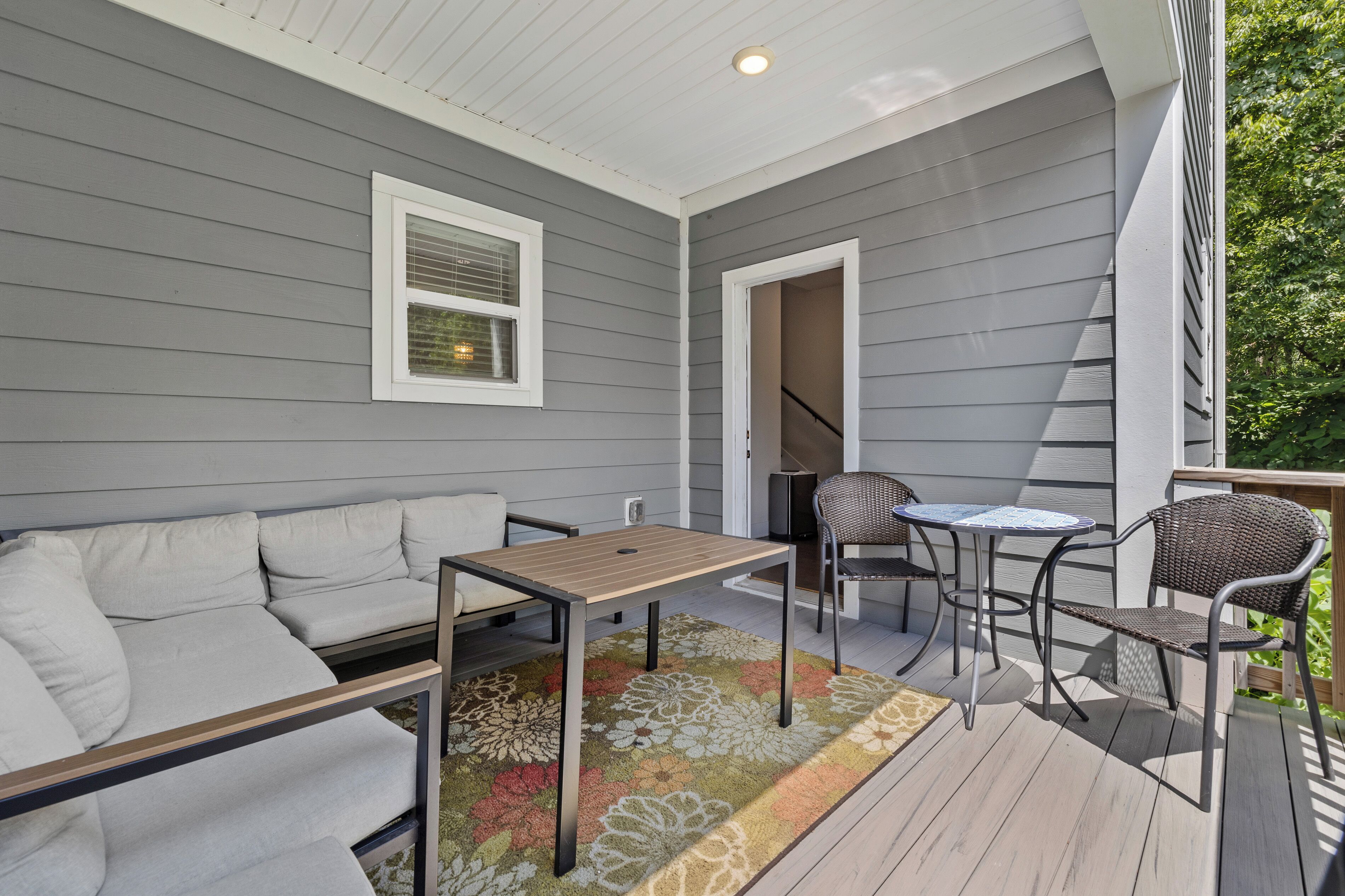 Un patio cubierto con una pared exterior gris, una ventana con marco blanco, un sofá, una mesa y dos sillas.
