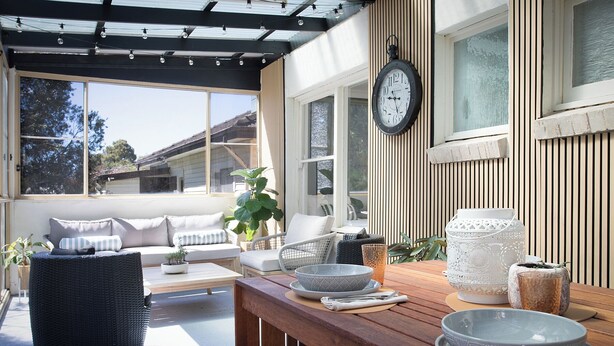 A covered outdoor seating area with a wooden bench, a table with bowls and a candle, and a clock on the wall. - Highland Heart Cottage, Newly Renovated, Spacious & Family Friendly (Bowral)
