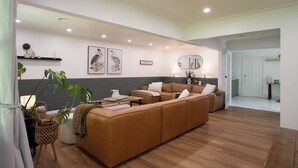 A spacious living room with a large brown sofa, a coffee table, and a side table. There are two wall-mounted picture frames, a mirror, and a door leading to another room. - Highland Heart Cottage, Newly Renovated, Spacious & Family Friendly (Bowral)