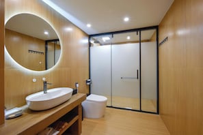 Un baño moderno con tocador de madera, un espejo ovalado grande, un lavamanos blanco y una zona de ducha con mampara de vidrio.