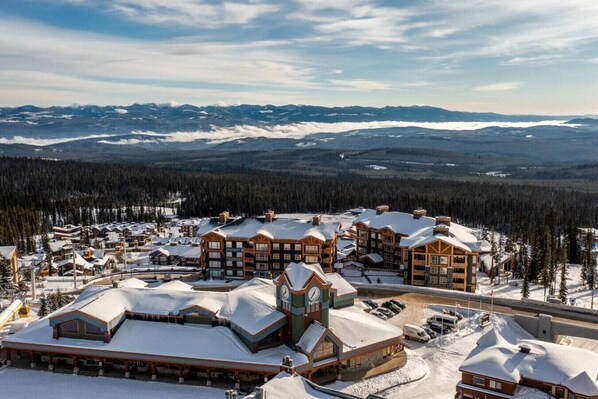 A snow-covered resort with multiple wooden buildings, a clock tower, and a view of snow-covered mountains in the background.