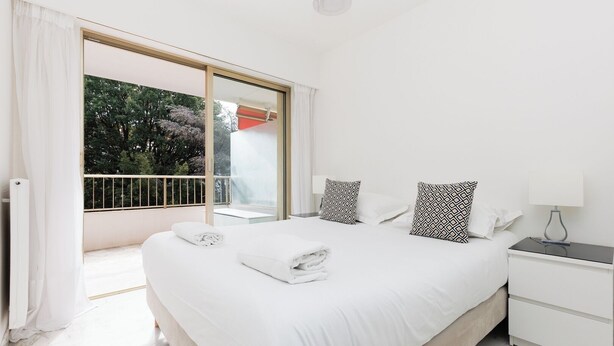 Un dormitorio con una cama grande, dos almohadas estampadas, una mesita de noche con lámpara y vistas a la vegetación a través de un ventanal corredero.