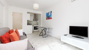 Una sala moderna con un sofá gris, cojines rojos y blancos, un mueble blanco para el televisor y una zona de comedor con mesa y sillas.