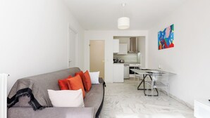 Una sala moderna con un sofá gris, cojines rojos y blancos, una mesa de comedor transparente con sillas, y una cocina al fondo.