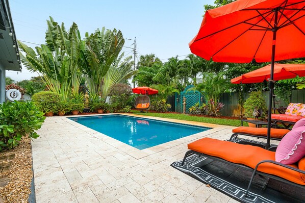 Une piscine dont le fond est recouvert de carrelage, entourée d’une végétation luxuriante et agrémentée de mobilier d’extérieur, notamment de chaises longues orange et de parasols rouges.
