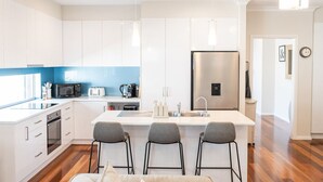 A modern kitchen with white cabinets, a stainless steel refrigerator, a microwave, a toaster, and a sink. There are three bar stools and a window providing natural light. - The Lake House—Spacious Group Haven by the Bay (Victor Harbor)