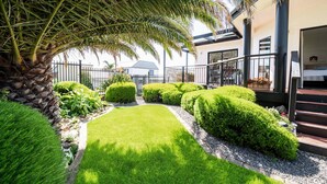 A well-maintained garden with a palm tree, neatly trimmed bushes, and a clear pathway leading to a building with a balcony. - BaeView: Panoramic Group Stay Above Encounter Bay (Encounter Bay)