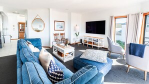 A modern living room with a blue sofa, a white coffee table, and a grey armchair. There is a large flat-screen TV mounted on the wall, a wooden cabinet, and a round mirror. - BaeView: Panoramic Group Stay Above Encounter Bay (Encounter Bay)