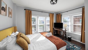 Una habitación de hotel con cama, escritorio con televisor y ventanas grandes con cortinas.