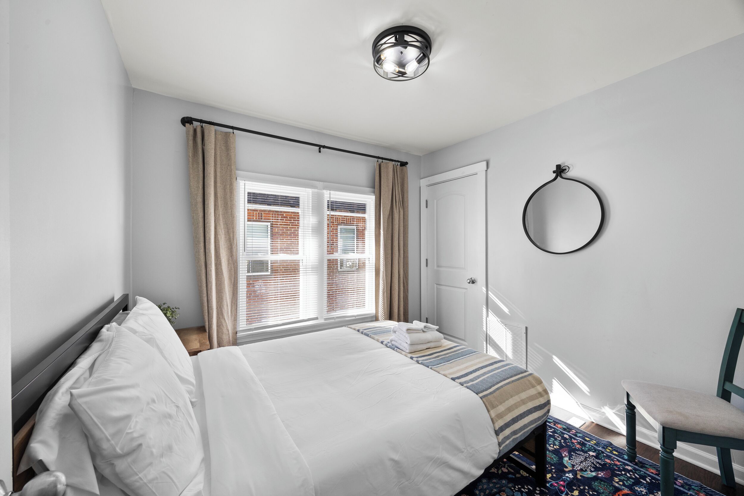 A bedroom with a bed, a window with curtains, a door, a chair, and a wall-mounted ring light.