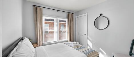 A bedroom with a bed, a window with curtains, a door, a chair, and a wall-mounted ring light.