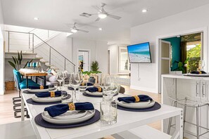 Un comedor con una larga mesa preparada para una comida, una escalera, un televisor y vistas al exterior.