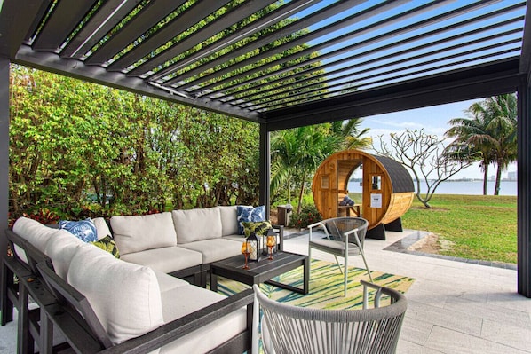 A modern outdoor seating area with a sofa, chairs, and a table. There is a wooden barrel structure and lush greenery in the background.