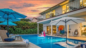 Ein Schwimmbad mit Liegestühlen und Sonnenschirmen, umgeben von einem angelegten Garten und einem zweistöckigen Gebäude.