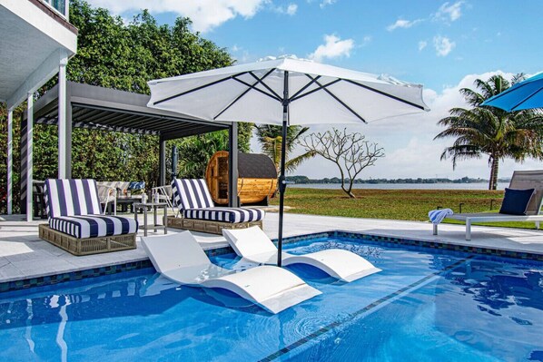 Piscina con sillones de descanso y sombrillas, rodeada por un porche cubierto.