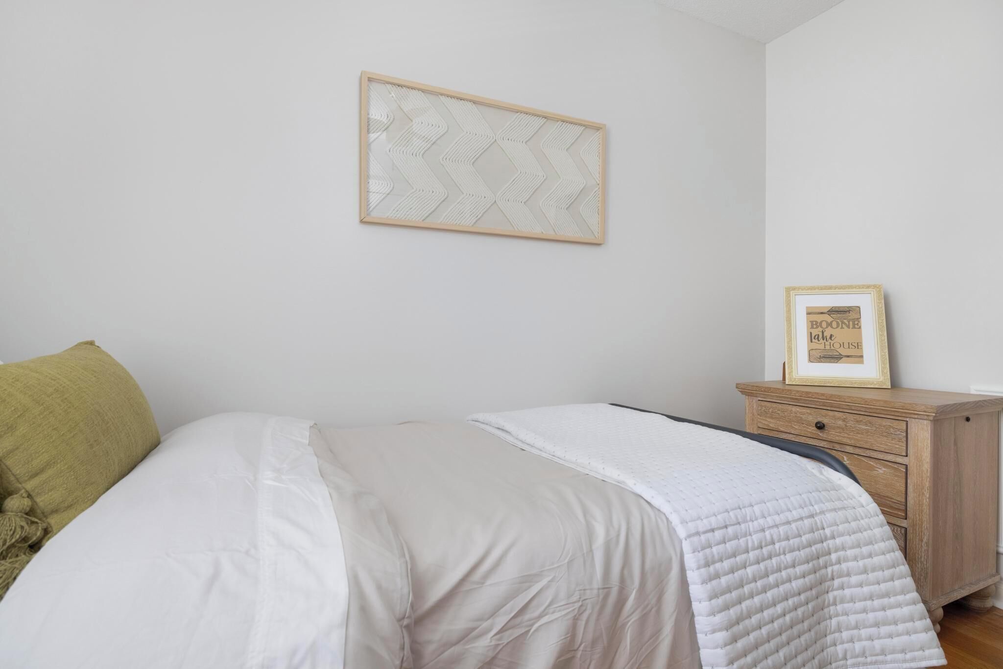 Ein ordentlich bezogenes Bett mit weißer Decke, ein Nachttisch aus Holz mit gerahmtem Bild und ein Wandbehang mit Muster.