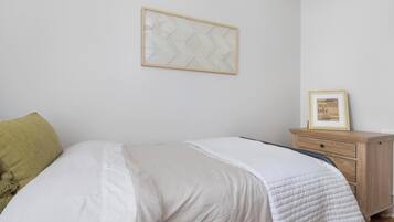 Ein ordentlich bezogenes Bett mit weißer Decke, ein Nachttisch aus Holz mit gerahmtem Bild und ein Wandbehang mit Muster.
