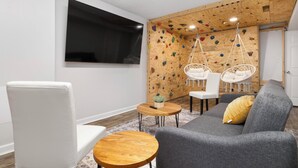 Ein modernes Wohnzimmer mit einem grauen Sofa, einem weißen Sessel und einem Holztisch. Im Hintergrund ist eine Kletterwand zu sehen.