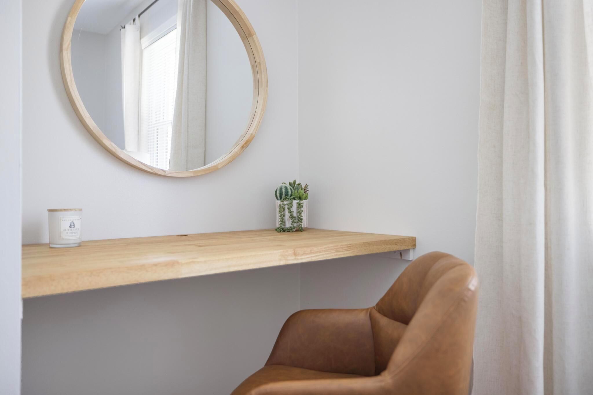 Une pièce avec une  étagère en bois, un miroir rond, une petite plante en pot et une chaise marron.