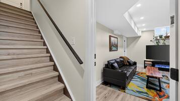 A modern living room with a staircase, a black leather sofa, a colorful rug, and a flat-screen TV.