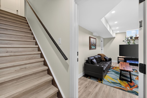 A modern living room with a staircase, a black leather sofa, a colorful rug, and a flat-screen TV. - Niagara Wellness Retreat at Awarenessome Haven (Thorold)