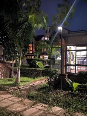 A nighttime outdoor area with a pathway, palm trees, and a building with large windows. - View Point Cottages (Ndhiwa)